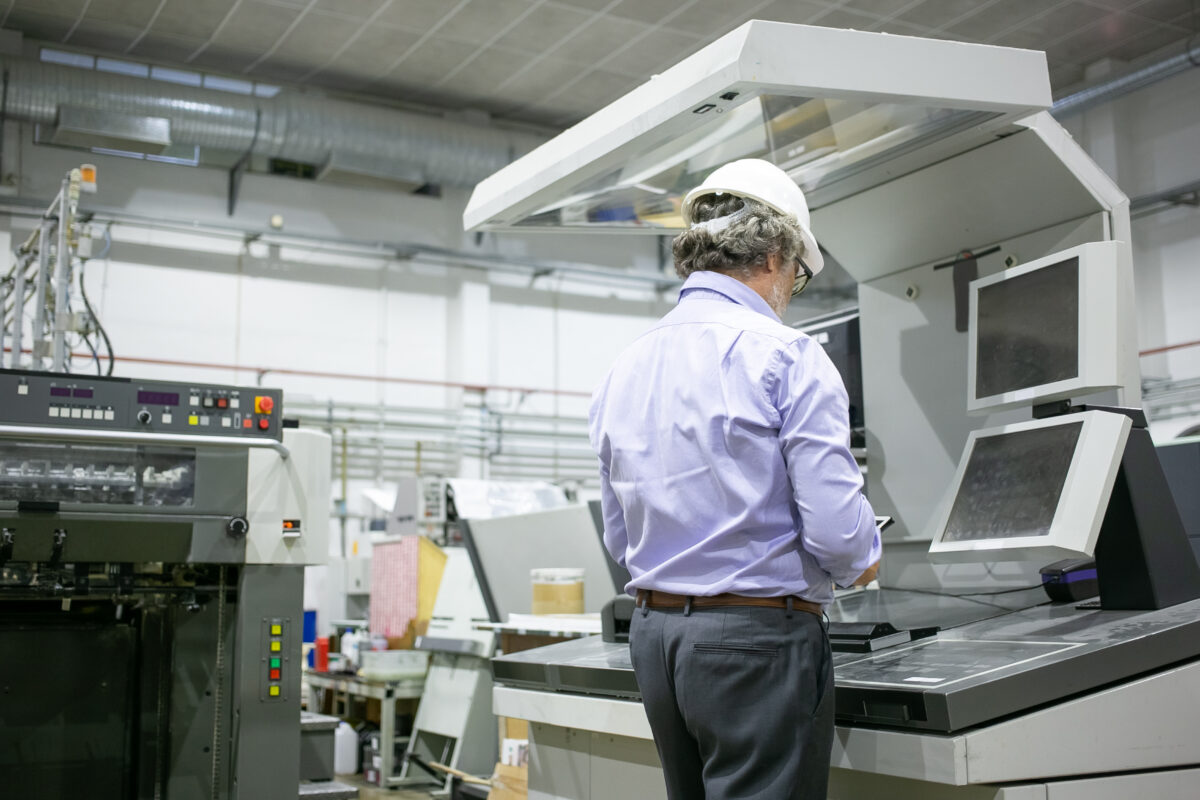 Male plant engineer in hardhat and glasses standing at industrial machine, using digital device.